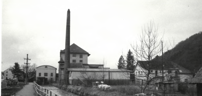 Das schwarz-weiße Bild zeigt eine Industrieanlage mit einem hohen Schornstein, umgeben von mehreren Gebäuden und karger Vegetation.