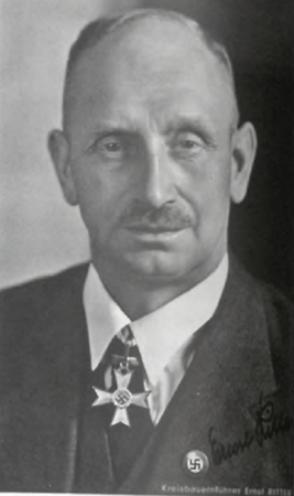 Ernst Ritter. Ein älterer Mann mit Glatze und Schnurrbart trägt einen Anzug mit einem Eisernen Kreuz und einer NSDAP-Parteiabzeichen-Nadel.