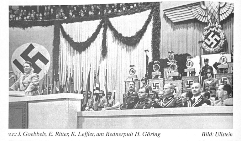 Parteiversammlung der NSDAP: Eine Versammlung in einem dekorierten Saal mit nationalsozialistischen Symbolen, wo eine Person an einem Podium spricht.