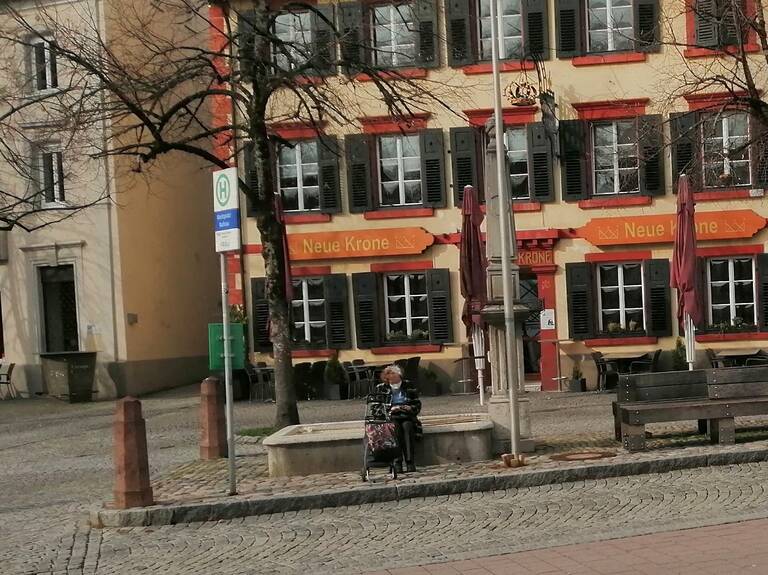 Kronenbrunnen in Alt-Schopfheim: Eine ältere Frau sitzt auf einer Bank vor einem gelben Gebäude mit roten Fensterrahmen und dem Schriftzug "Neue Krone" unter grauem Himmel.