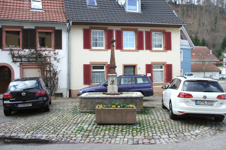 Aubrunnen in Alt-Schopfheim: Vor einem Haus mit roten Fensterläden steht ein Brunnen auf einem Kopfsteinpflasterplatz, flankiert von einem schwarzen und einem weißen Auto.