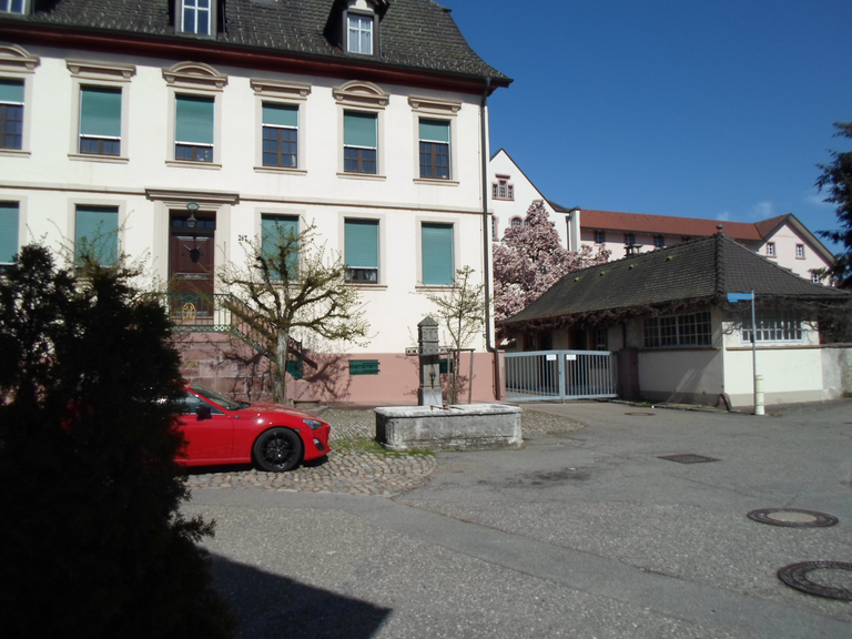 Klingelebrunnen / Krafftbrunnen: Ein rotes Auto steht vor einem großen historischen Gebäude mit grünen Fensterläden und einem angrenzenden Torhaus, umgeben von blühenden Bäumen.