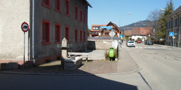 Hammerschmied-Brunnen. Ein ruhiger Straßenabschnitt mit einem Gebäude, einem Verbotsschild für Lkw, einem Brunnen und mehreren geparkten Autos im Hintergrund.