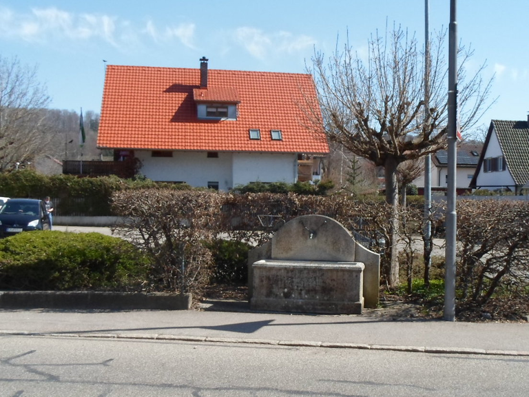 Hieberbrunnen. Ein Haus mit rotem Ziegeldach ist hinter einer Hecke und einem steinernen Brunnen am Straßenrand zu sehen.
