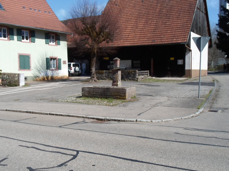 Löwenbrunnen. Ein kleiner Brunnen steht auf einem Platz vor einem Gebäude mit roten Dachziegeln und grünen Fensterläden, daneben befindet sich eine Straße mit einem Verkehrsschild.