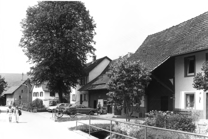 Foto von Ingrid Schubert: Enkenstein 1983. Eine ländliche Straße mit mehreren traditionellen Häusern, geparkten Autos und zwei Spaziergängern im Vordergrund.