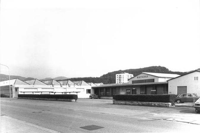 Foto von Ingrid Schubert: Fahrnau 1983. Das Bild zeigt eine industrielle Gebäudestruktur mit einem langgestreckten Flachdach und dem Schriftzug "Rietschle" auf einer der Fassaden, daneben sind parkende Autos und Hügel im Hintergrund zu sehen.