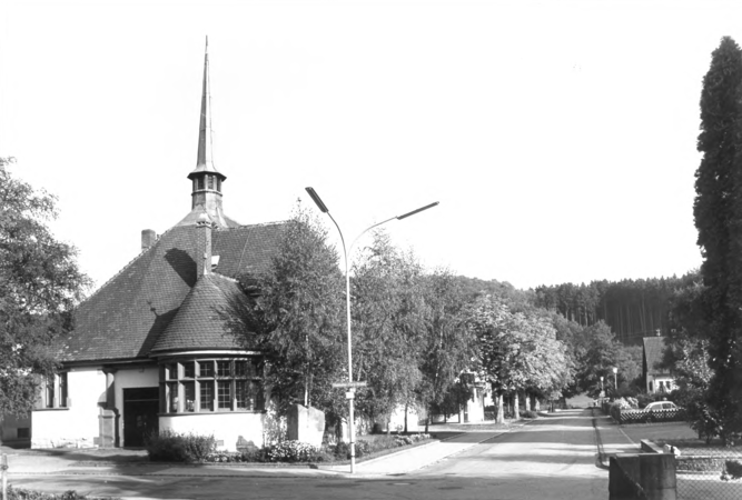 Foto von Ingrid Schubert: Fahrnau 1983. Ein Gebäude mit einem spitzen Turm steht an einer von Bäumen gesäumten Straße bei Tageslicht.