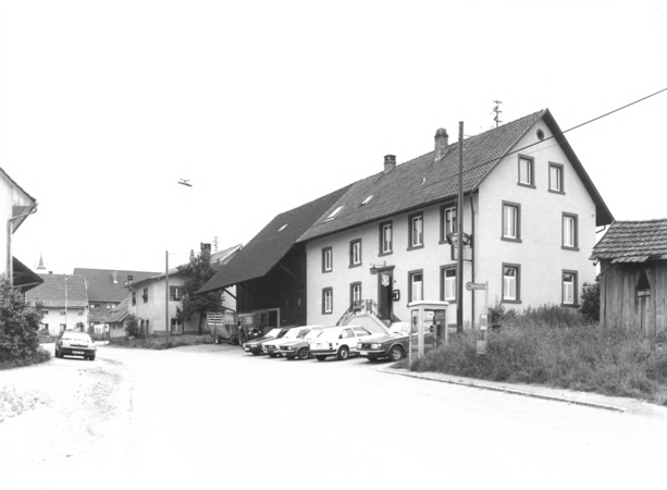 Foto von Ingrid Schubert: Kürnberg 1983. Ein großes, traditionelles Haus steht an einer ländlichen Straße mit mehreren parkenden Autos davor und einem kleinen Flugzeug am Himmel.