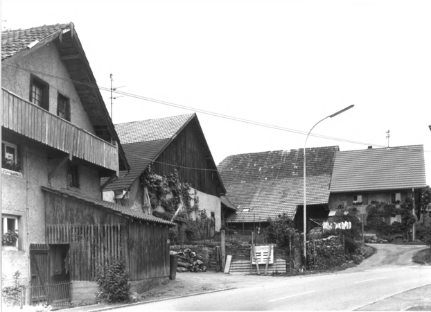 Foto von Ingrid Schubert: Kürnberg 1983. Das Bild zeigt eine ländliche Straßenecke mit mehreren traditionellen Häusern mit steilen Dächern und Holzelementen.