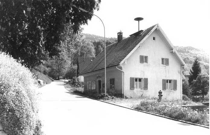 Foto von Ingrid Schubert: Raitbach 1983. Ein weißes Haus mit mehreren Fenstern und Fensterläden steht neben einer kurvigen Landstraße, umgeben von Bäumen und Hügeln.