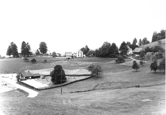 Foto von Ingrid Schubert: Schweigmatt 1983. Ein Freibad in einer ländlichen Landschaft mit Wiesen, mit verstreuten Bäumen und mehreren Gebäuden auf einem Hügel.