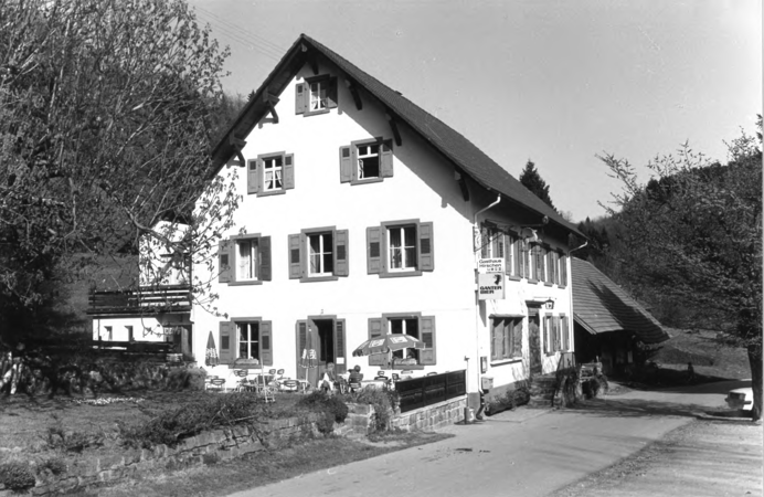 Foto von Ingrid Schubert: Raitbach 1983. Ein traditionelles, dreistöckiges Gebäude mit Schindeldach und Fensterläden steht in einer ländlichen, bewaldeten Umgebung neben einer Straße; davor sind Tische und Sonnenschirme aufgestellt.