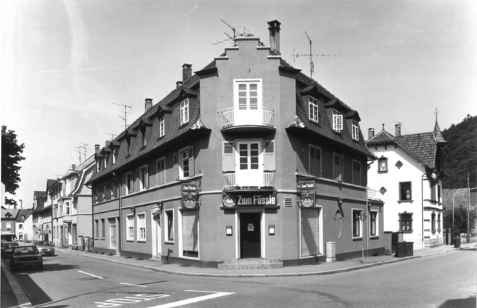 Foto von Ingrid Schubert: Schopfheim 1983. Ein dreistöckiges, traditionelles Gebäude mit dem Namen "Zum Fässle" an einer Straßenecke in einer ruhigen Wohngegend.