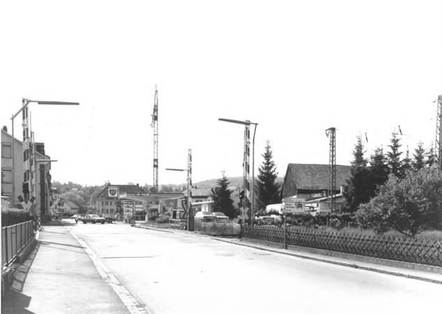 Foto von Ingrid Schubert: Schopfheim 1983. Eine Straße mit einer Eisenbahnkreuzung und einem Tankstellenbereich, umgeben von Gebäuden und Bäumen.
