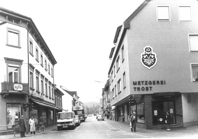 Foto von Ingrid Schubert: Schopfheim 1983. Schwarz-weiß-Fotografie einer Straßenszene mit mehreren Gebäuden, darunter eine Metzgerei mit der Aufschrift "Metzgerei Trost", und mehreren geparkten Autos.