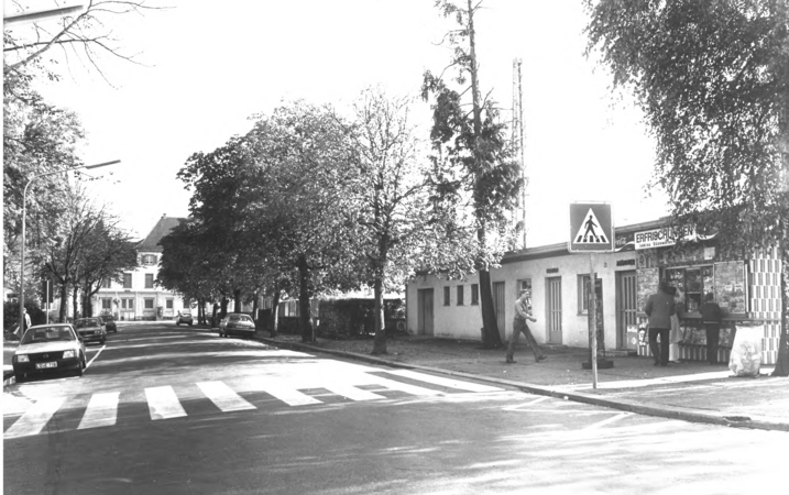 Foto von Ingrid Schubert: Schopfheim 1983. Eine Straße mit Zebrastreifen führt an einem Kiosk vorbei, während mehrere Menschen und Autos zu sehen sind, umgeben von Bäumen und Gebäuden.
