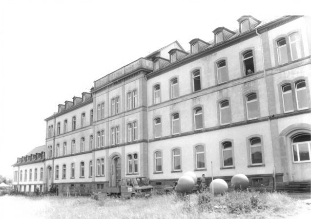 Foto von Ingrid Schubert: Wiechs 1983. Ein großes, mehrstöckiges, historisches Gebäude mit mehreren Fenstern und einem LKW sowie drei großen Kugeln im Vordergrund.