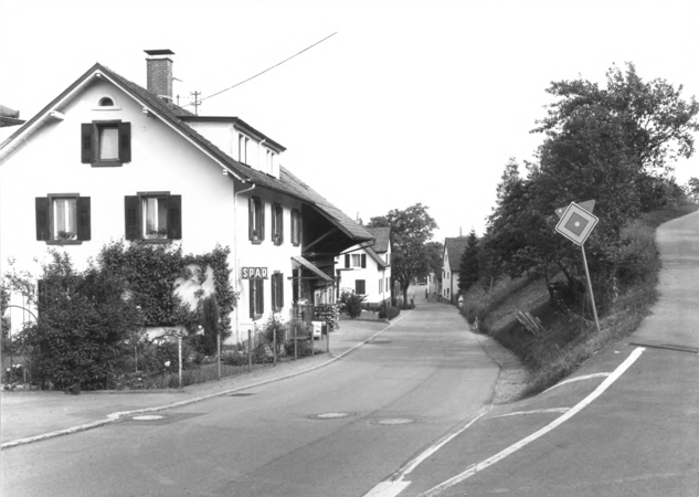 Foto von Ingrid Schubert: Wiechs 1983. Eine Schwarz-Weiß-Aufnahme zeigt eine ländliche Straße mit Häusern, einem "SPAR"-Laden und einem Verkehrsschild, umgeben von Bäumen und Vegetation.